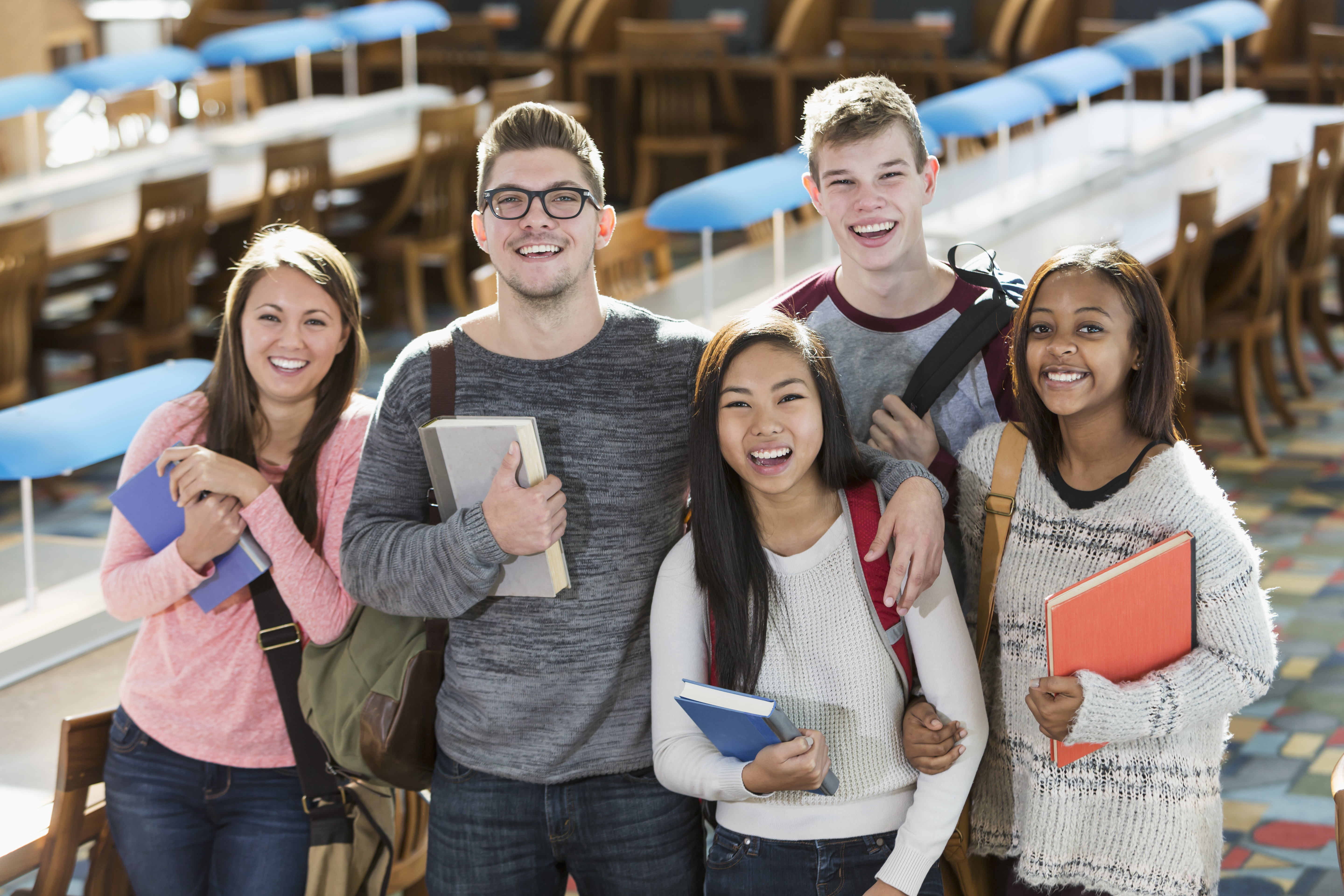 Portrait of a multiracial group of high school or college students in a library reading room, carrying book bags and textbooks, smiling at the camera. There are two young men and three young women standing together in the foreground. Behind them are rows of tables and wooden chairs with blue reading lamps.